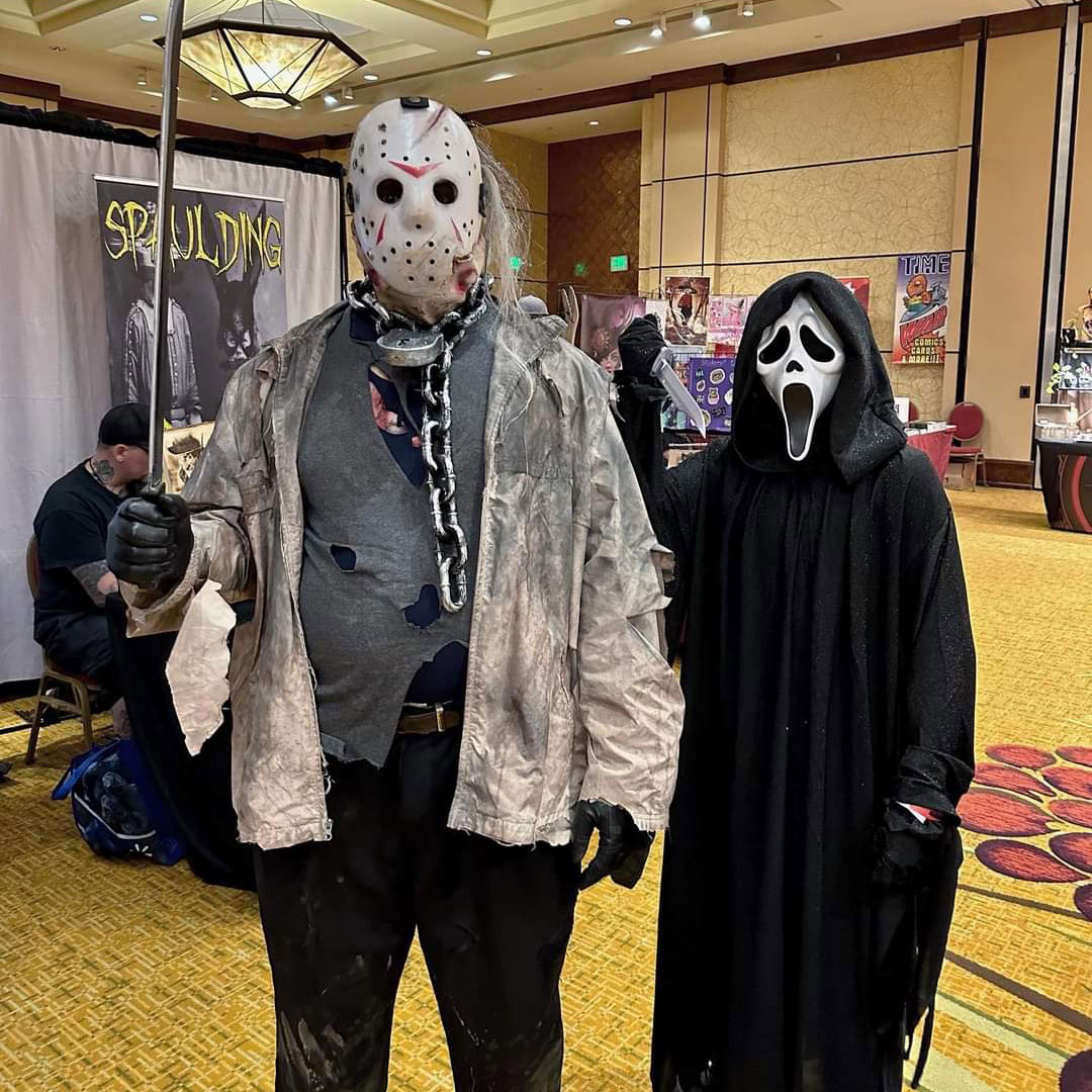 Person dressed in a ghostface, white mask with mouth open and eyes dropping and a bloack robe with a fake knife, standing nec to a man in a Jason mask with white hair sticking out, plastic chain around neck, dirty white longsleeve and ripped shirts undernearth with black pants and black gloves on