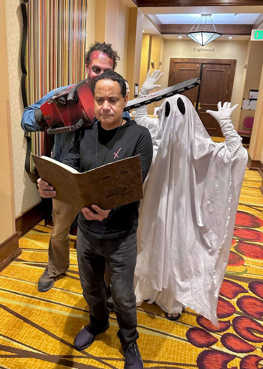 A dark skinned man in black with black (Jeffery Riddick), holding a brown book with a man Ash from Army of Darkness, a chainsaw hand blood on him with dark spiky hair and a Lil Boo, a ghost standing behind the man with the book in the hotel hallway 