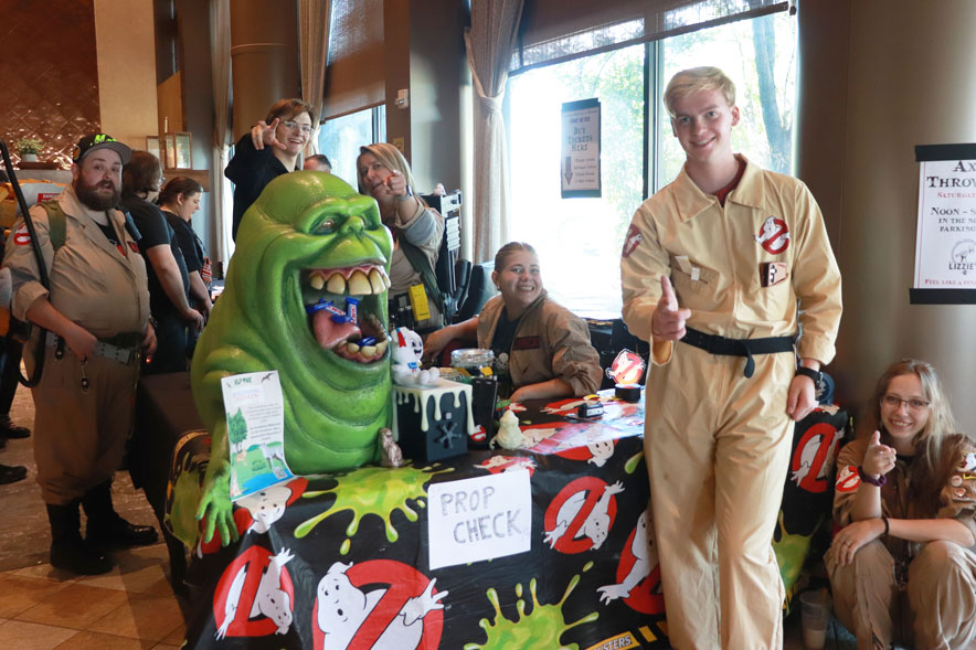 A Ghostbuster with pink hair, in a beigh flight suit with warning tape all over taking a picture of a silent killer doctor who is bald in a messy white coat and blue scrub pants, while another female blonde Ghostbuster in the same outfit is looking at a vendor booth full of plants