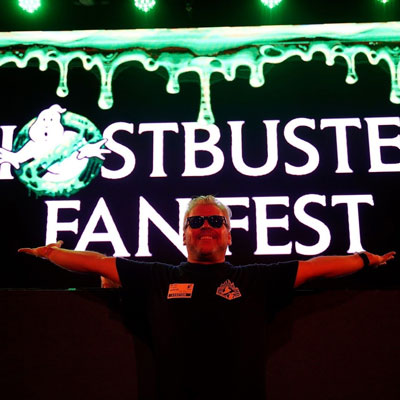 Smurf, an extremely sexy blonde smiling man with glasses and in a Colorado Ghostbusters tshirt, arms spread wide in front of a Ghostbusters Fanfest convention sign.